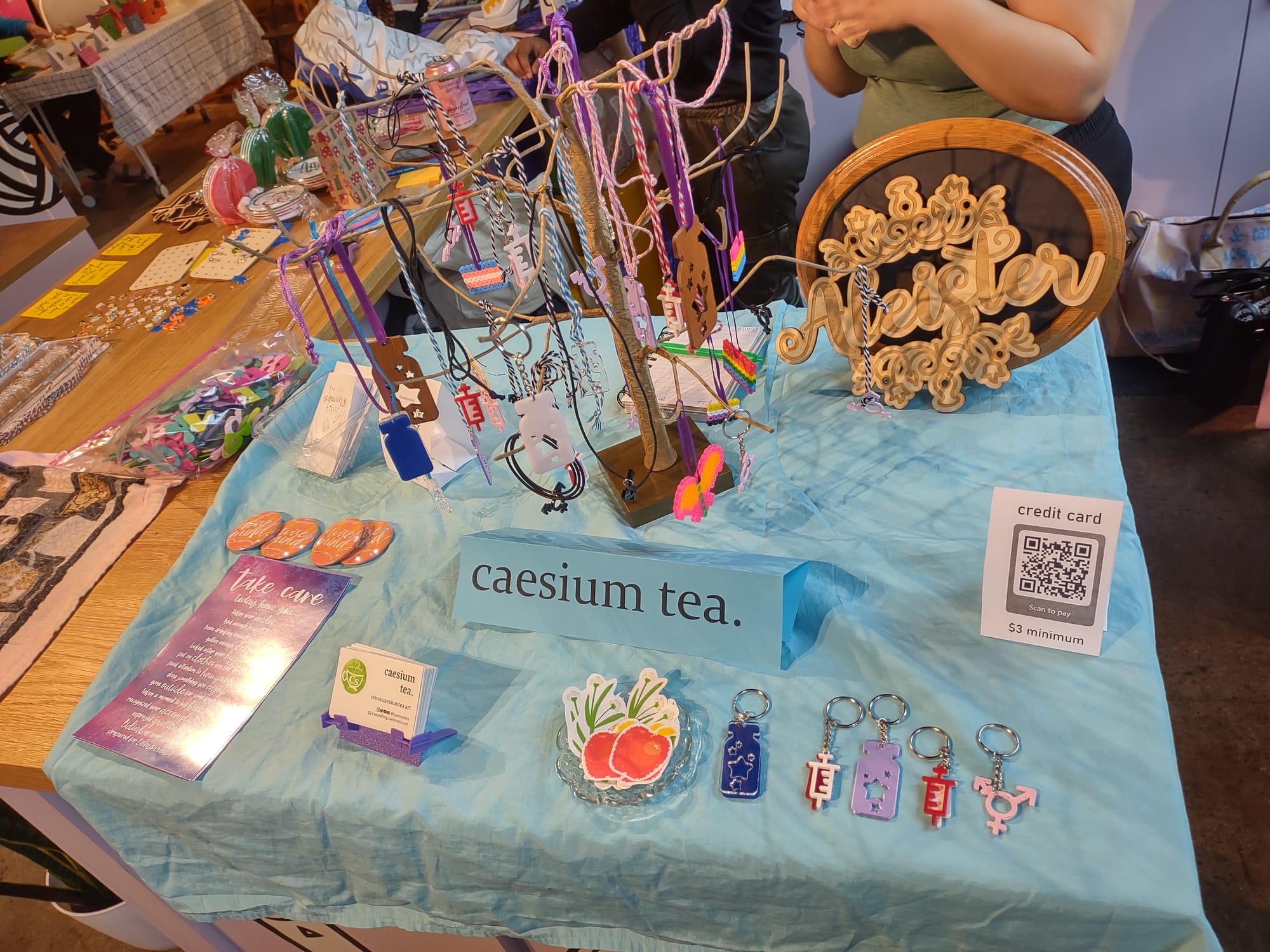 A craft fair table display. There's a light blue sheet over the table and a folded piece of paper labeled "caesium tea" in the center. Several items from the above images are scattered around the table, with necklaces and ornaments hanging from a small tree.