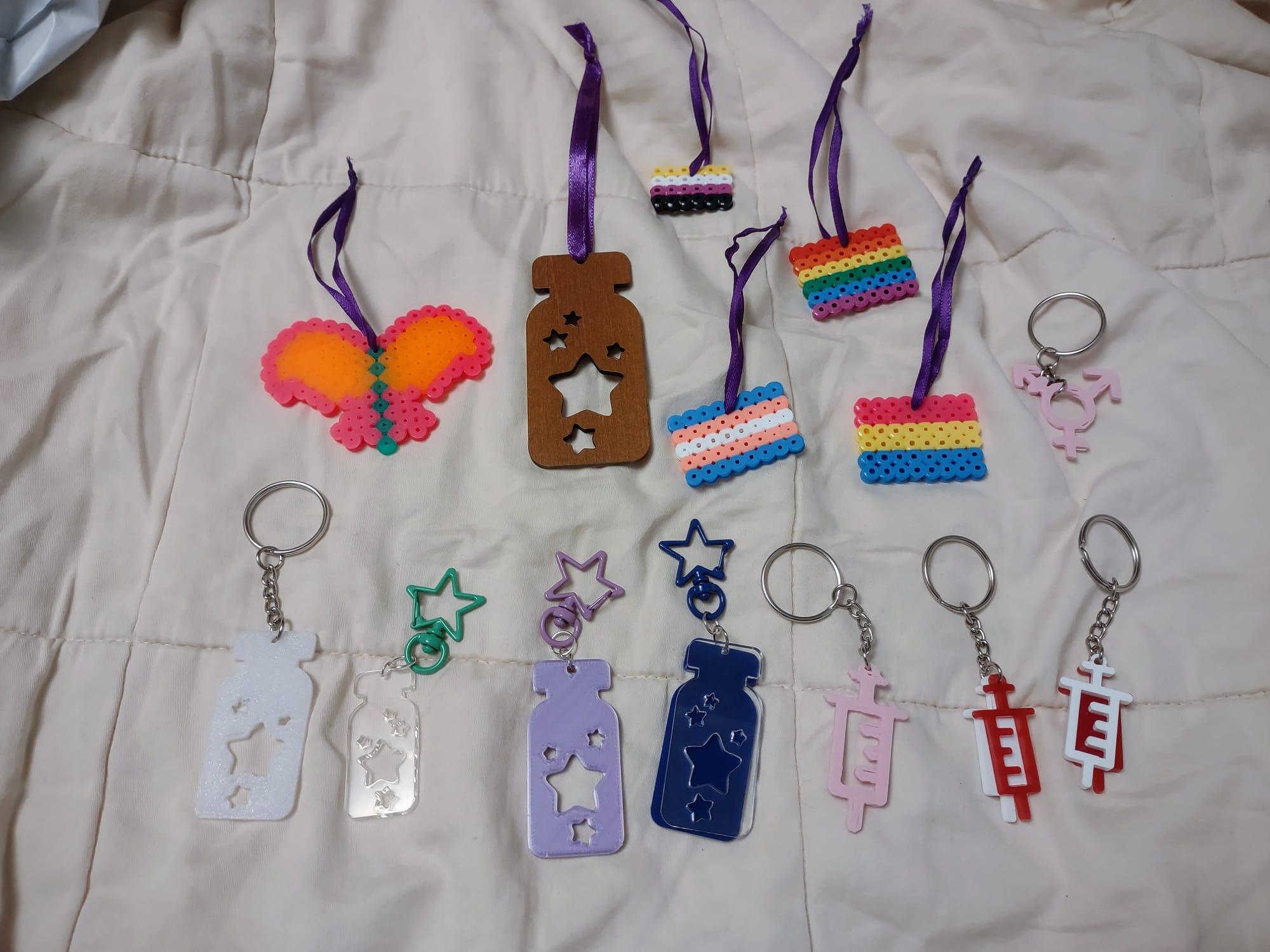 A variety of keychains and ornaments laid out on a blanket. Some are syringe designs, some are medicine vials with a star shape cut out inside, one is a trans symbol, and some are pride flags made of Perler beads.