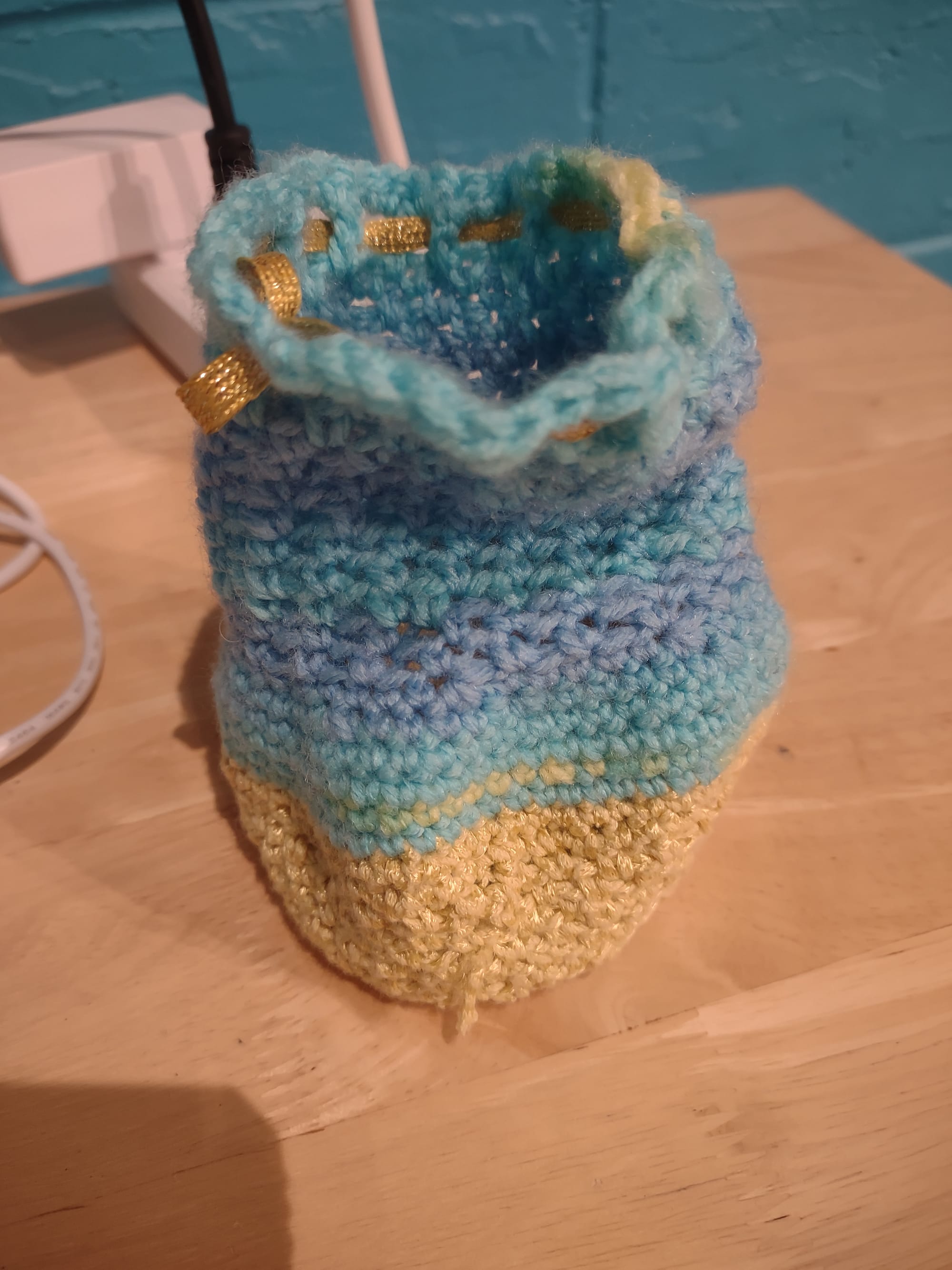 A pouch made of shiny yellow yarn at the bottom and then the rest in a yarn that's a gradient of light blue tones with a few yellow spots. There's a gold cord strung through a series of holes at the top to make a drawstring. It's sitting on a wooden table next to a power strip.