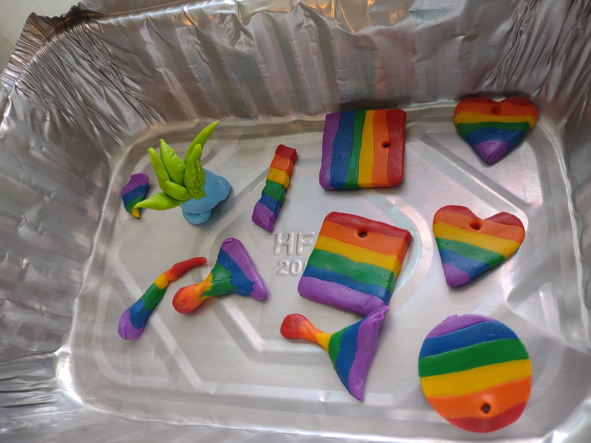 An aluminum pan holding a variety of polymer clay charms. Most are various flat shapes in rainbow stripes with holes at the top for hanging. There's also a little figurine of the Pokemon Oddish.