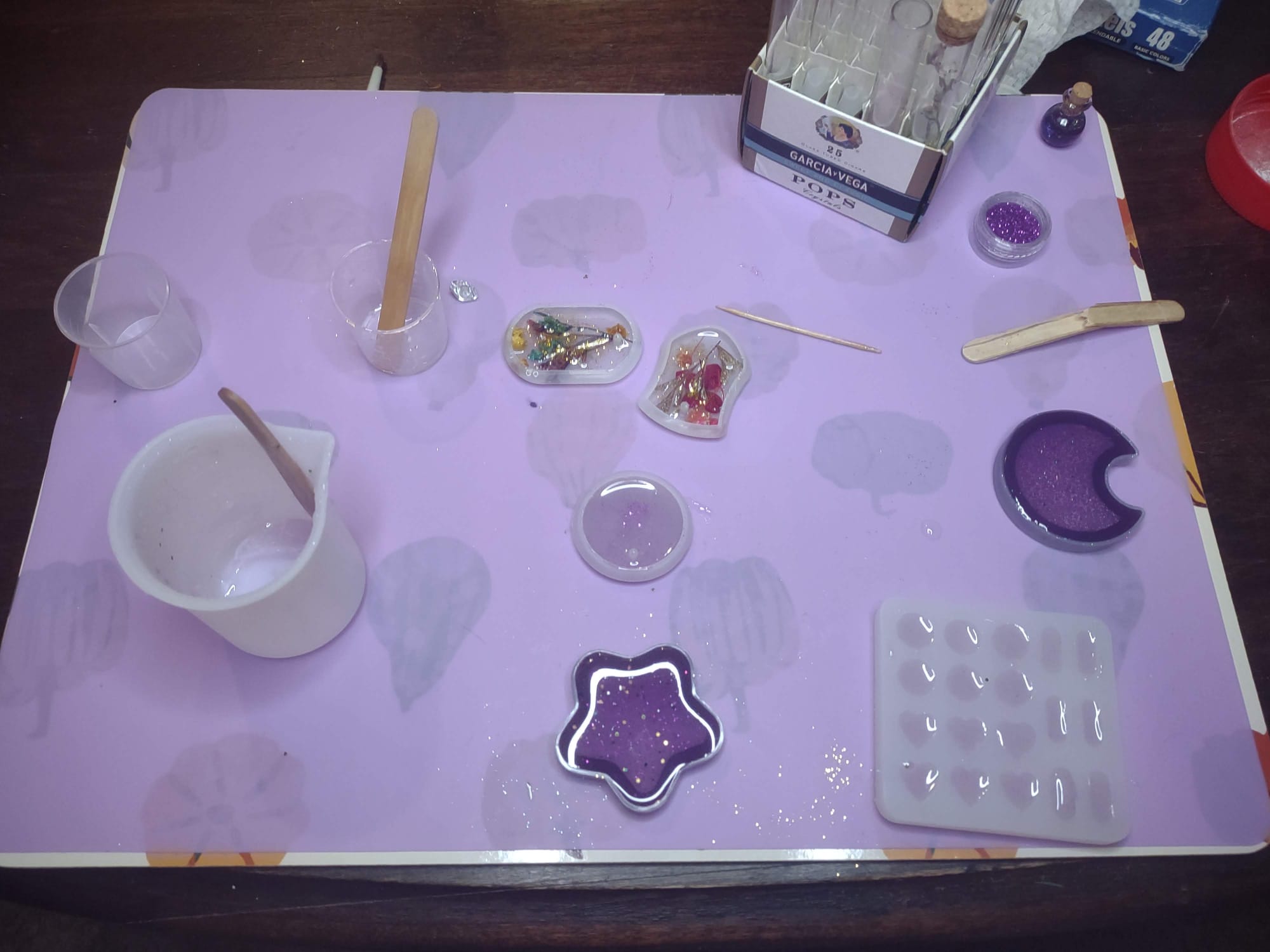 A variety of silicone molds spread out on a silicone mat, along with cups of clear resin containing popsicle stick stirrers. The molds include a star and moon with purple resin and some pendants with clear resin and colorful flowers inside.