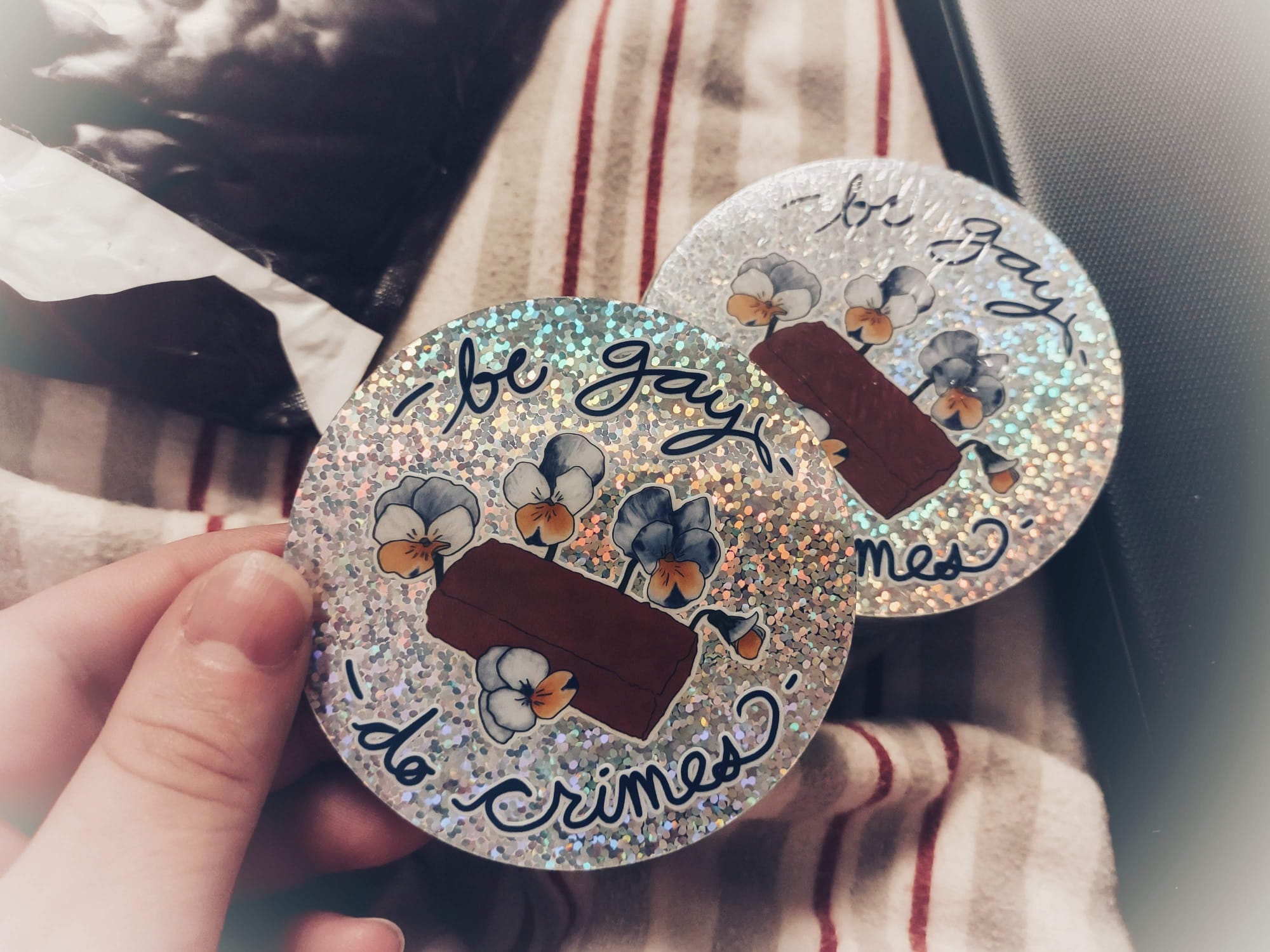 A hand holding a 3 inch round sticker with the words "be gay do crime" and an image of a brick surrounded by pansies on a glittery silver background.