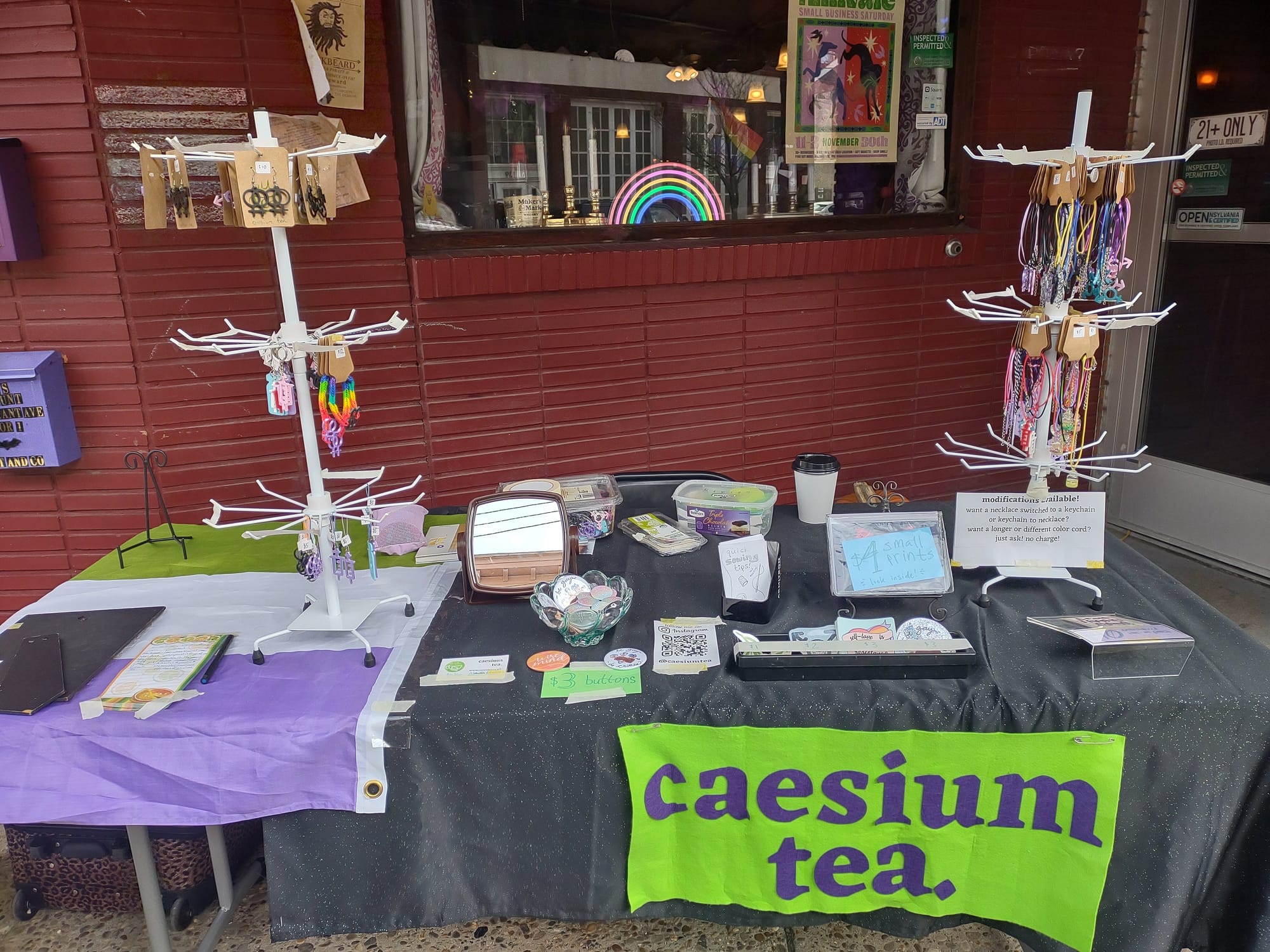 A table outside of a red brick building with a neon rainbow in the window. The table has a green and purple banner that says "caesium tea" on the front. On top of the table there are two white spoked towers with necklaces and charms hanging from the spokes.