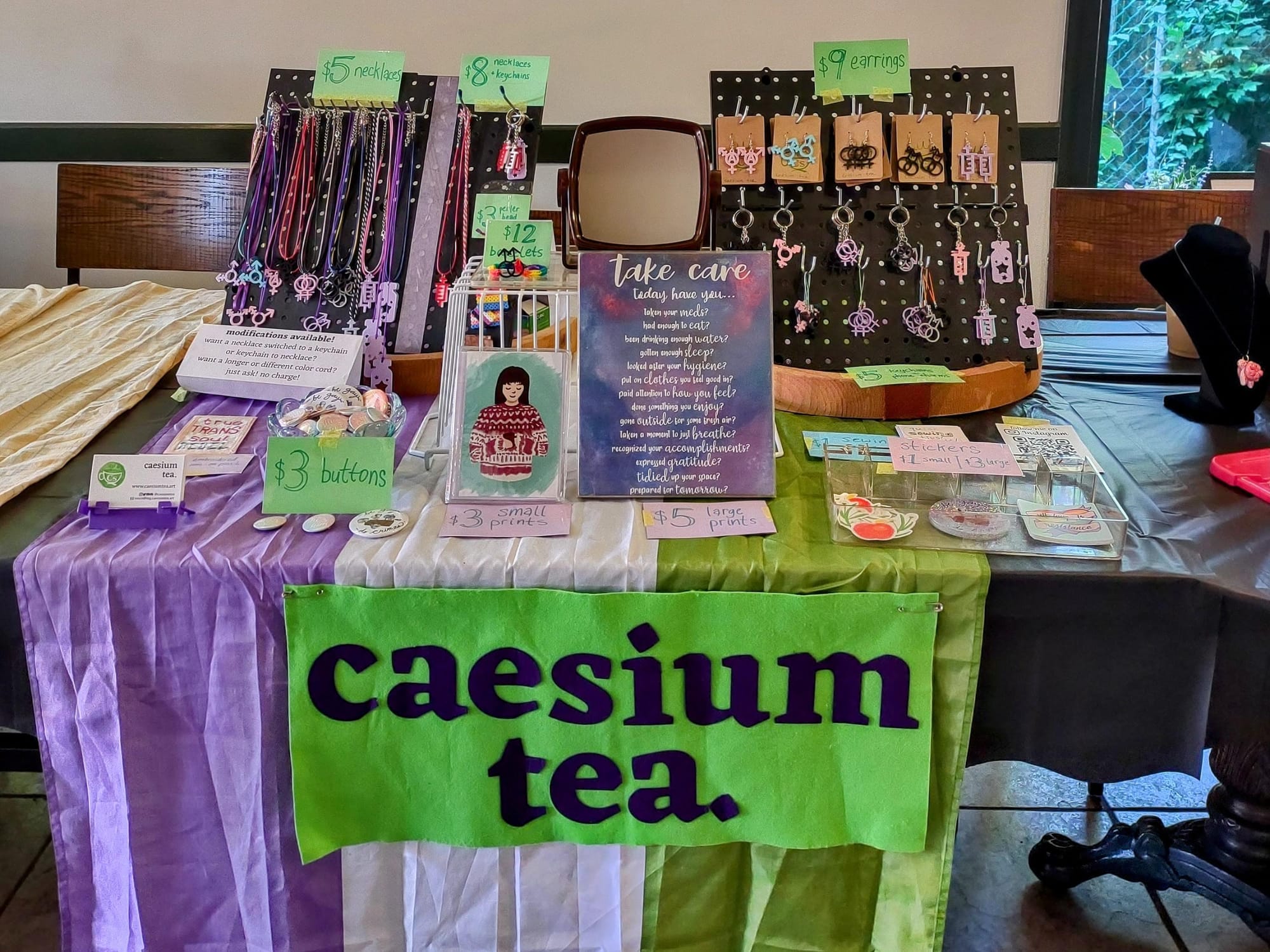 A table display at a craft fair with a green and purple sign on the front that says "caesium tea". There is a genderqueer pride flag spread over the table. On the table, there are two black pegboards with necklaces and keychains hanging from them, a poster that says "take care" above a list, a bowl of round buttons, and a smaller print of a painting of a person in a pink and white sweater holding a mug.