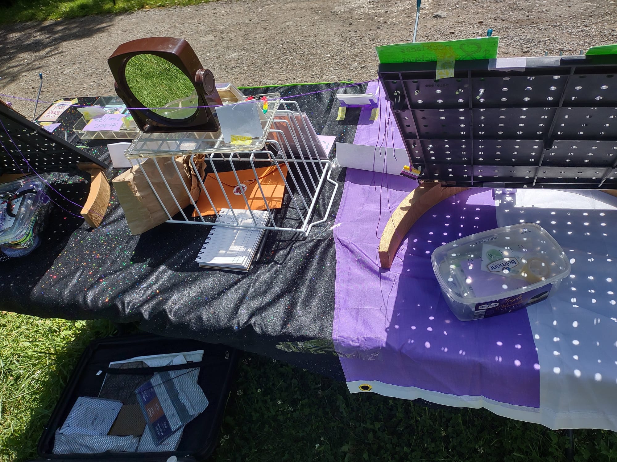 The table on the grass from above, seen from the back. The backs of the two black pegboards are visible, as well as a tupperware container, notebook, envelope, and brown paper bag on the table. There is an open suitcase on the ground behind the table.