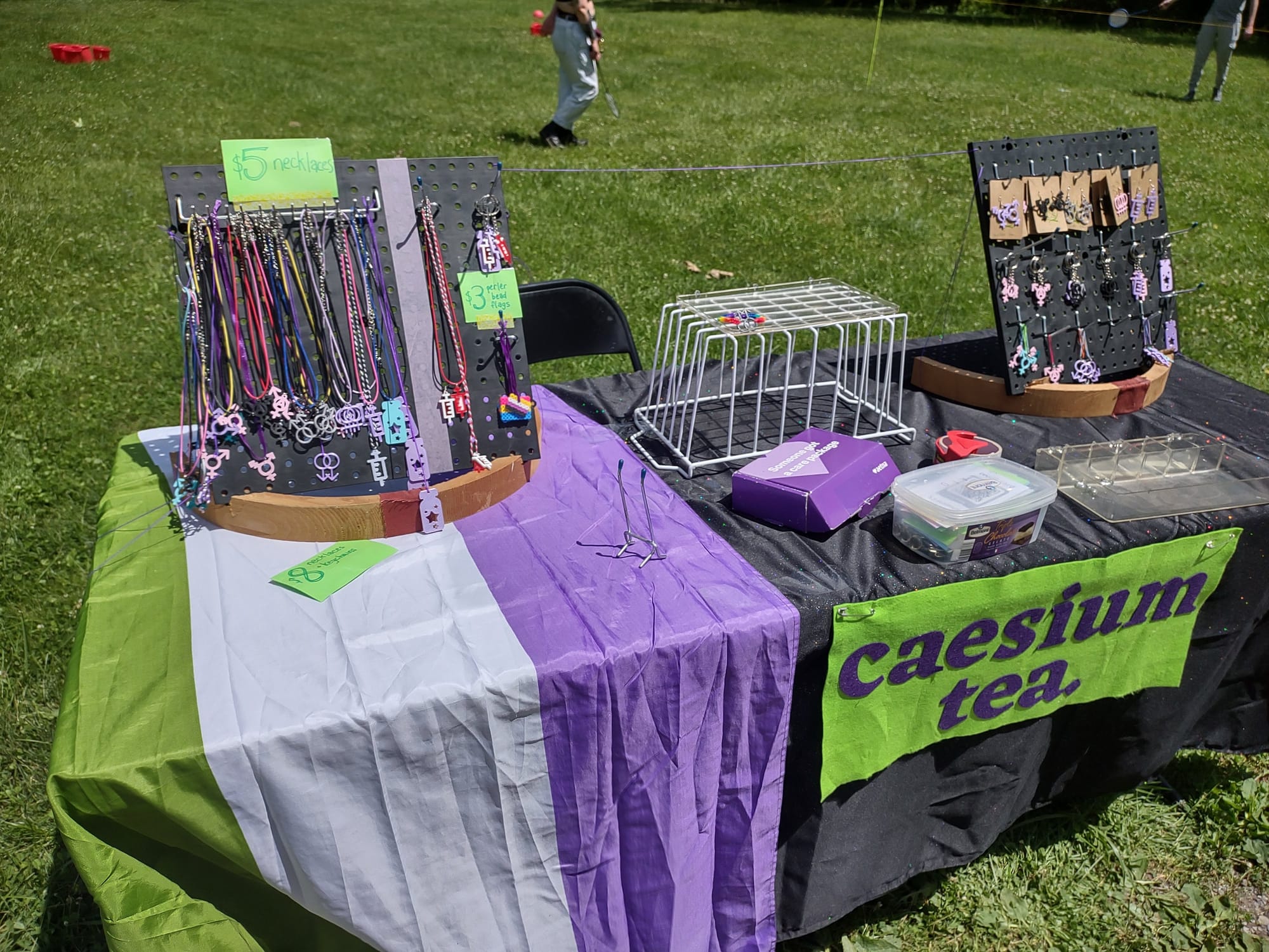 A vendor table similar to the one seen above, but it's only partially set up, and is outdoors on a patch of grass. There's the same genderqueer pride flag, "caesium tea" banner, and two black pegboards full of necklaces and charms. There are also a few other small boxes and tape scattered around the table.