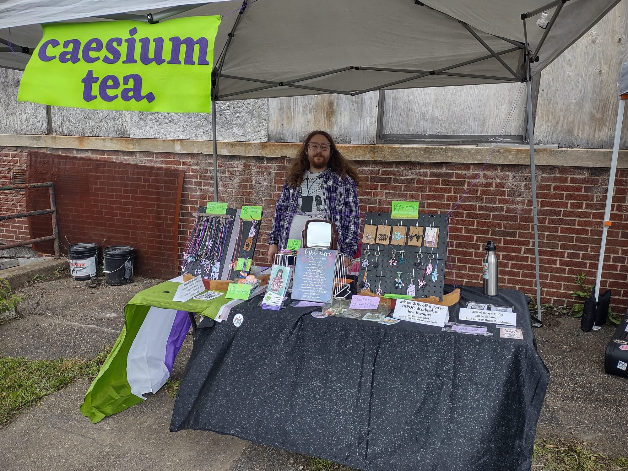 Vance standing behind a similar table setup to the ones seen above. This time there is a canopy tent over the table and the "caesium tea" banner is hanging from the tent at the top of the image. Vance is a white adult with long curly brown hair and a beard and mustache, wearing glasses and a purple plaid shirt, and smiling slightly to the camera. 