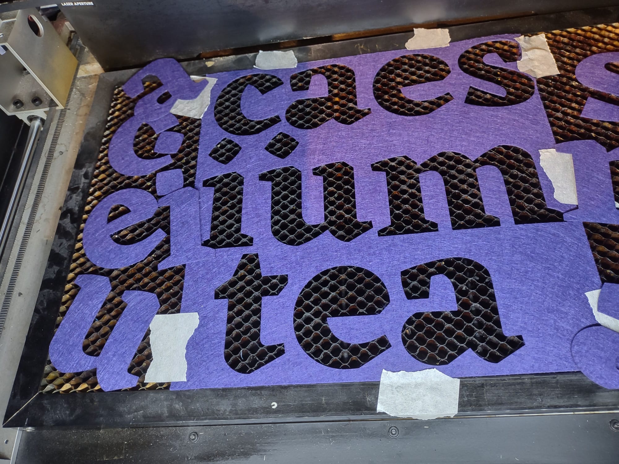 A piece of purple felt taped down to a honeycomb bed inside a laser cutter, with the letters of "caesium tea" cut out of it.