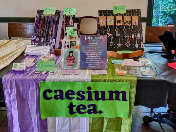 A table display at a craft fair with a sign on the front that says "caesium tea". There are necklaces, keychains, and prints.